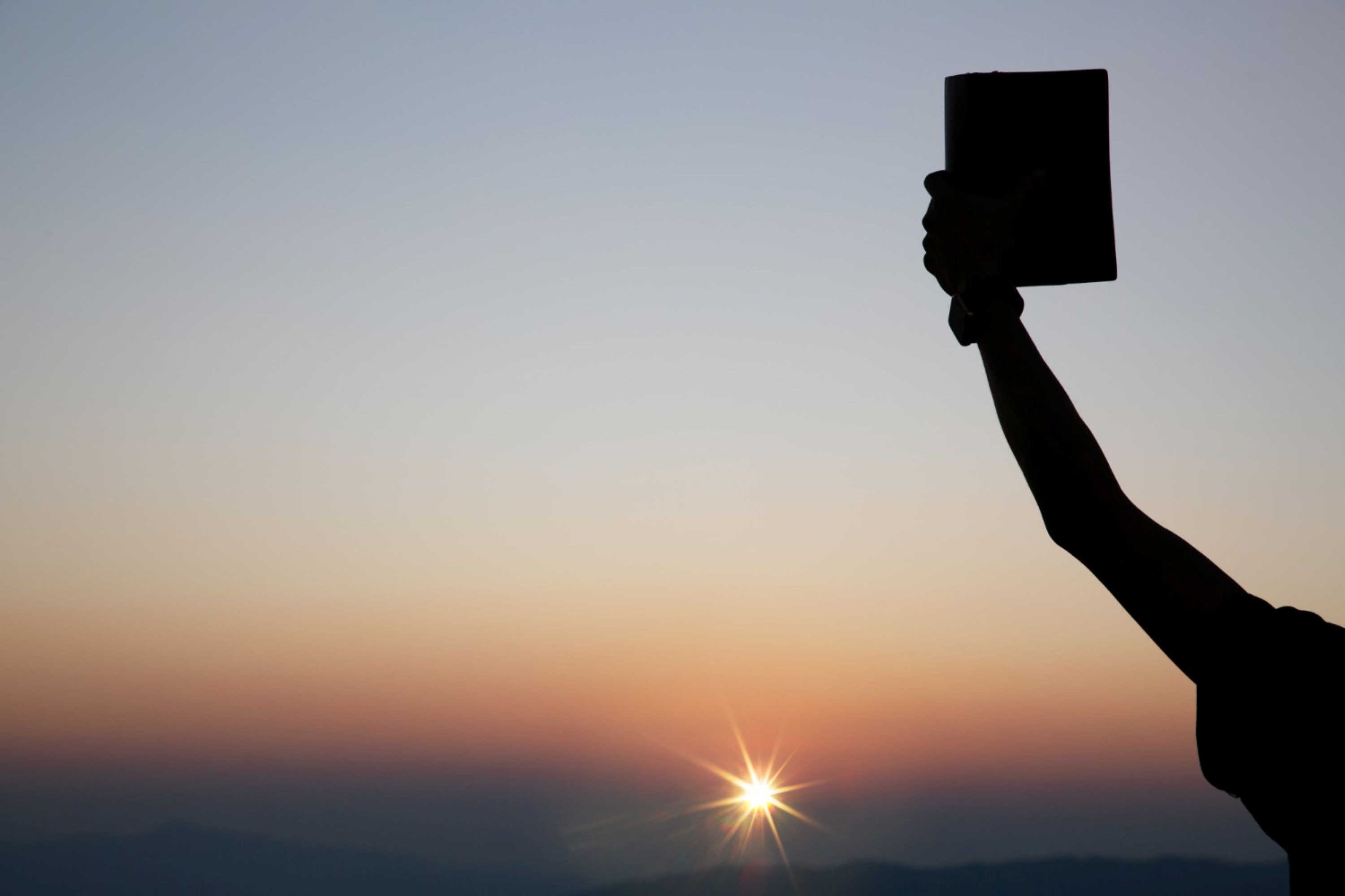 A person lifting up the Bible into the air as the sun sets.